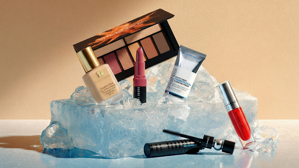 Winter makeup products including eyeshadow palette, foundation, lipstick, primer, mascara, and liquid lipstick displayed on ice