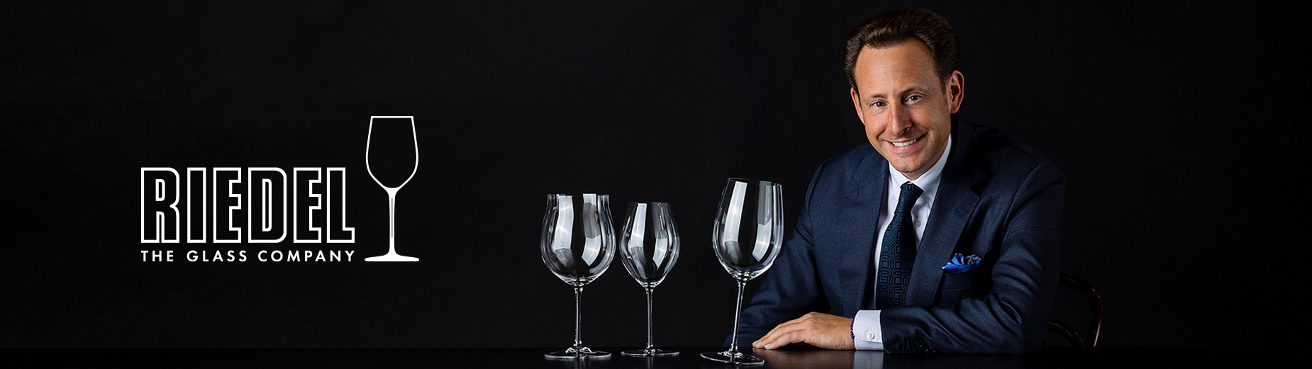 Man in suit with three elegant wine glasses and RIEDEL The Glass Company logo on black background